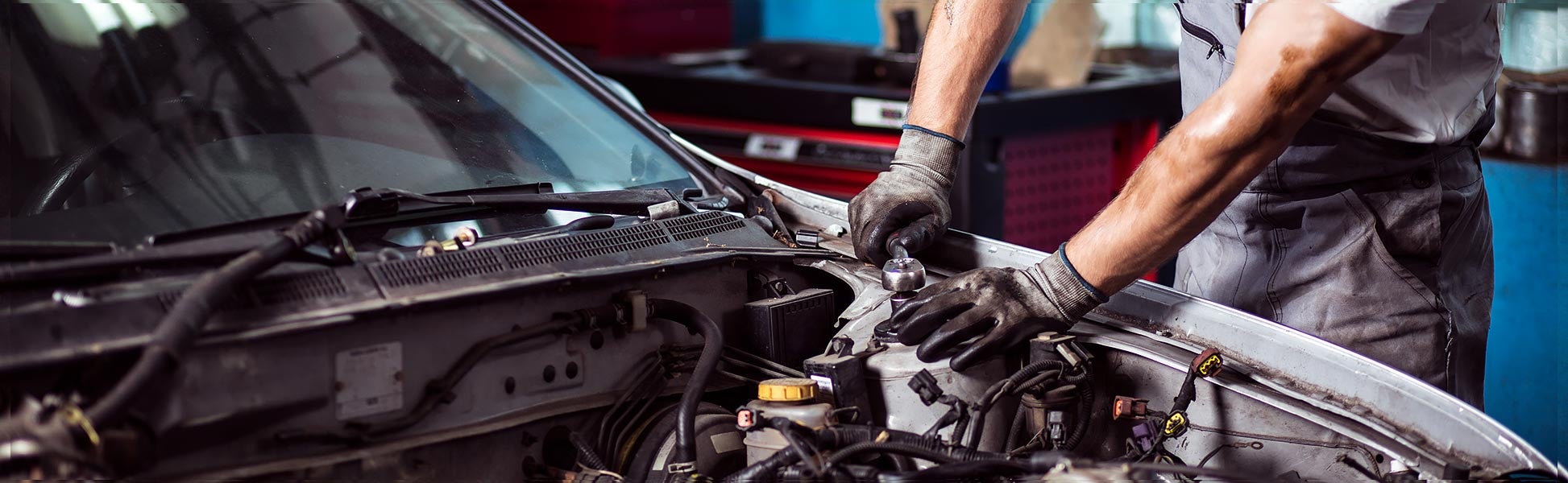 A mechanic, wearing work gloves, uses a wrench to repair a car engine under the open hood.