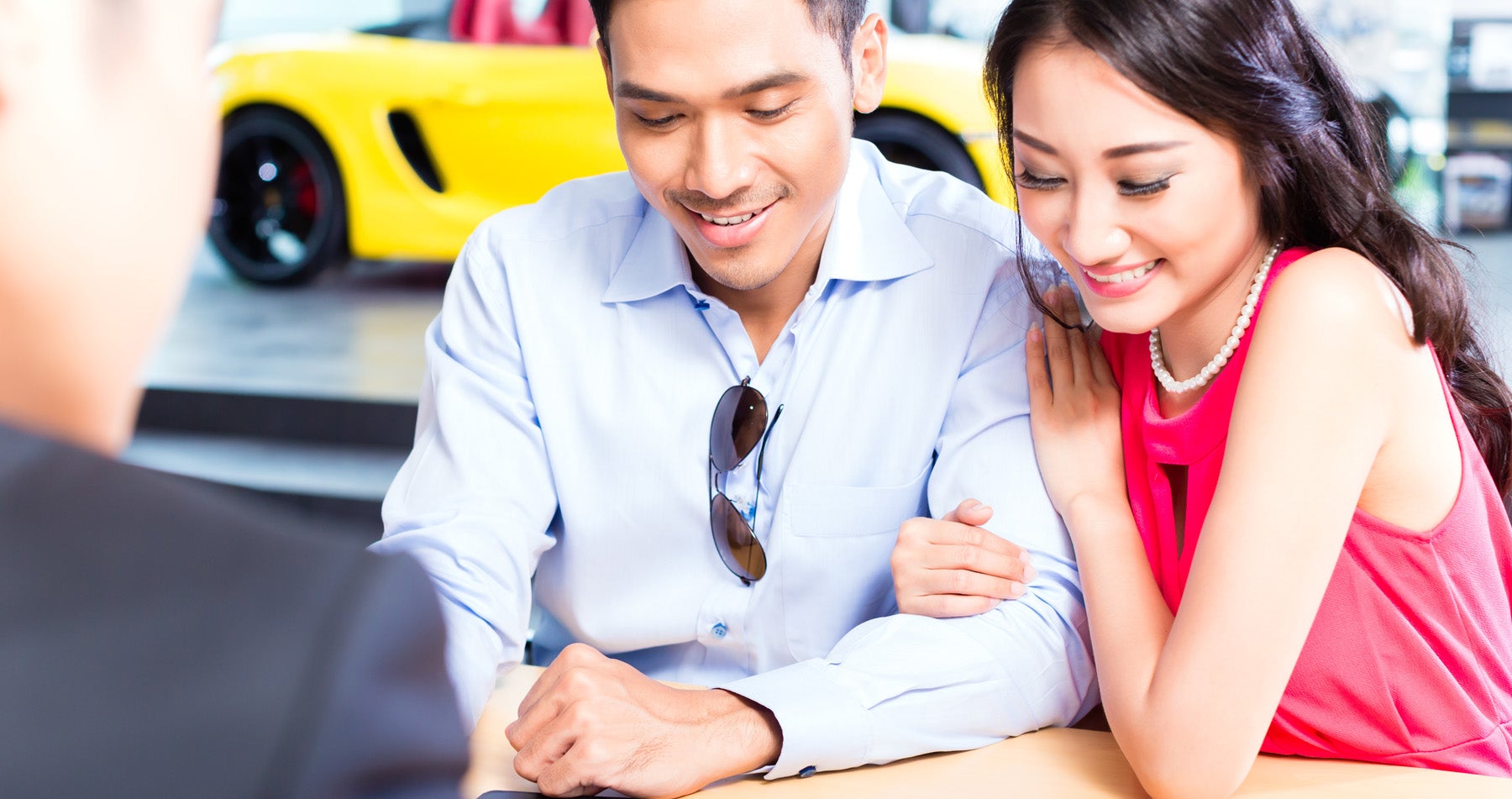 A smiling Asian couple looks at a document in front of them, consulting with a person whose back is visible. A yellow sports car is in the background.