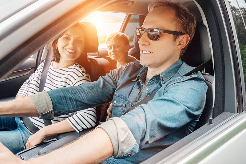 A family of three enjoying a car ride, all smiling, with sunlight entering the vehicle.