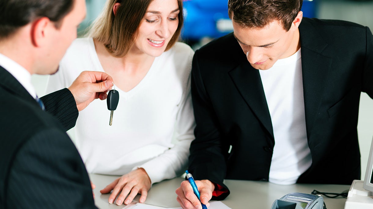 Salesperson handing car keys to a smiling woman as a man signs paperwork.