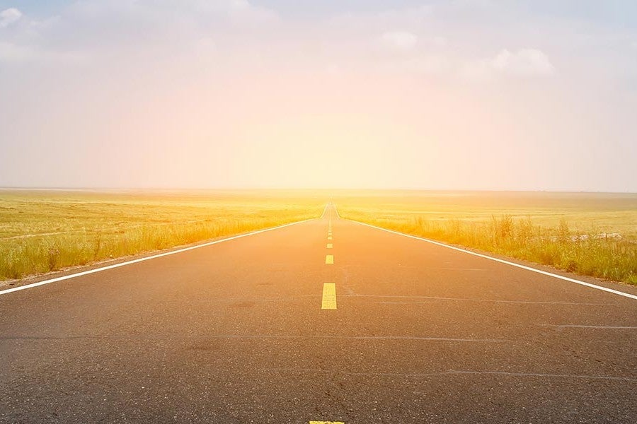 An empty paved road stretches into the distance, flanked by grassy fields under a hazy, bright sky.